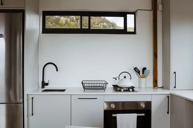 36m² 1-Bedroom Container House — Insulated Standard Configuration — supplier gallery view 7; Fitted container house kitchen with white cabinets, black tap, gas hob, oven and stainless fridge beneath a horizontal window