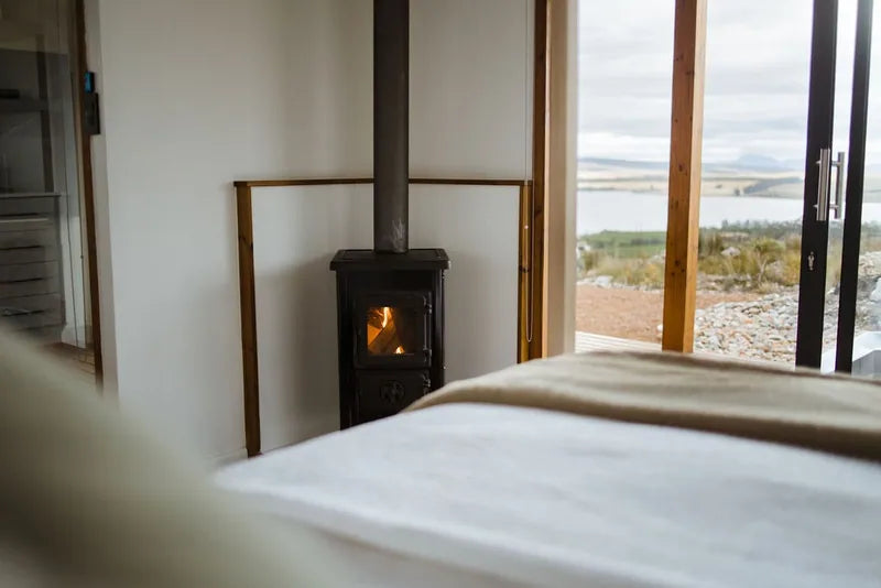 36m² 1-Bedroom Container House — Insulated Standard Configuration — supplier gallery view 9; Container house bedroom with a made bed, compact wood-burning stove and glazed door opening towards the landscape