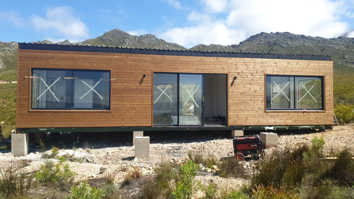 94m² 2-Bedroom Container House — Insulated Standard Configuration — supplier gallery view 6; Long timber-clad modular home with a run of black-framed windows, standing in open veld below mountains