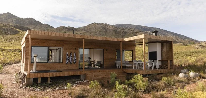 94m² 2-Bedroom Container House — Insulated Standard Configuration — supplier gallery view 1; Timber-clad modular home with a covered veranda and outdoor furniture, set in open veld with mountains behind