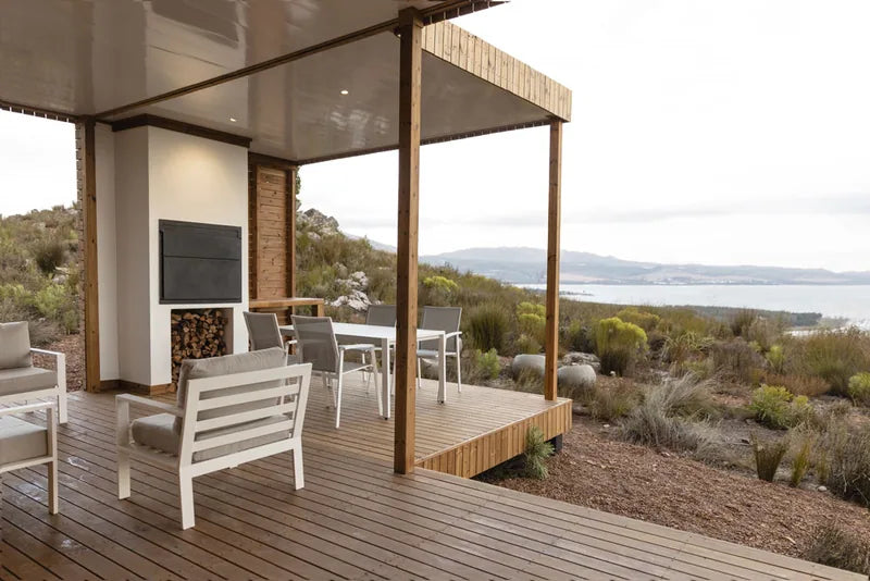 94m² 2-Bedroom Container House — Insulated Standard Configuration — supplier gallery view 2; Covered veranda with outdoor seating, a built-in fireplace and timber posts, looking across scrub to a lagoon